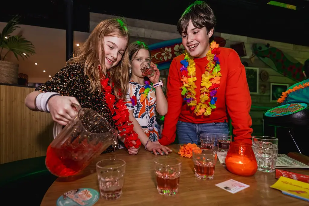 2 meisjes en een jongen staan rond een tafel. Meisje schenkt ranja in glaasjes.